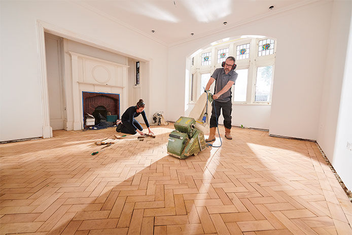 two people working to repair and restore a parquet period floor. The man is sanding the floor while the woman is cutting and repairing rotten floor boards
