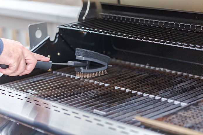 A close up image of a man's hand using a steel bristle brush to clean a gas burning grill