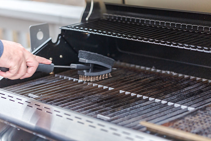 A person uses a black, long-handled grill brush to scrub the surface of the grates inside a stainless-steel propane grill.