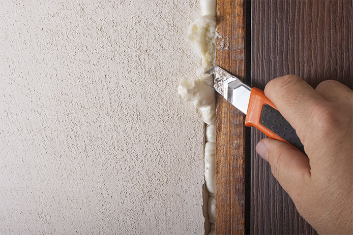 A close up image of a person using a orange utility knife to scultp down spray foam in a door frame