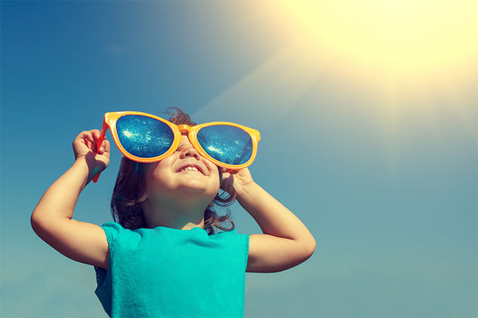 A little kid in a blue shirt holding up oversized sunglasses to their face. The sun is beaming down on the from the top right hand corner of the image 