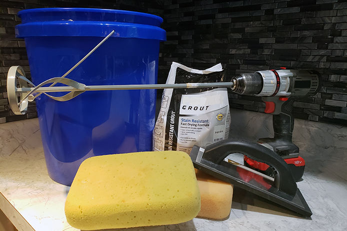 Supplies to apply tile backsplash. A blue five gallon bucket, grout, a drill and paint mixer, a yellow sponge and a grout float sitting on a countertop