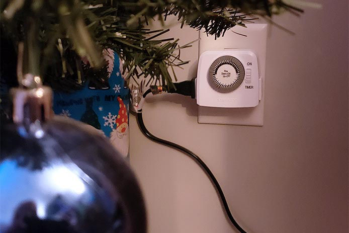 A household timer plugged in behind a Christmas tree with a blue ornament in the foreground
