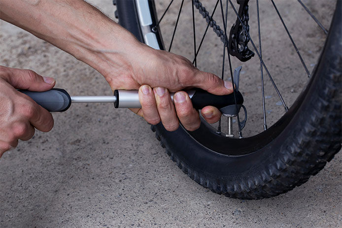 A hand is shown holding a tire gauge and checking the tire pressure of a bike tire.  The gauge is pressed firmly against the valve of the tire. The bike tire is a narrow, black road tire with a distinctive tread pattern.