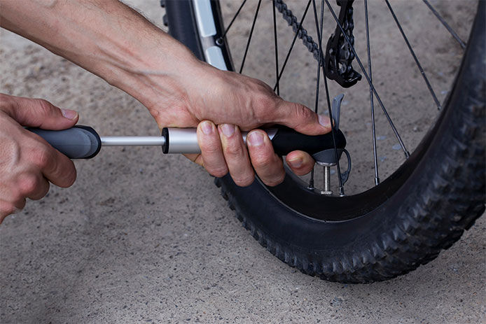 A hand is shown holding a tire gauge and checking the tire pressure of a bike tire. The gauge is pressed firmly against the valve of the tire. The bike tire is a narrow, black road tire with a distinctive tread pattern.