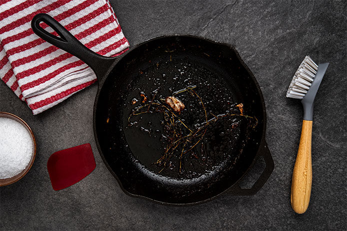 A scrubber brush laying on countertop next to a skillet that is dirty and has just been used