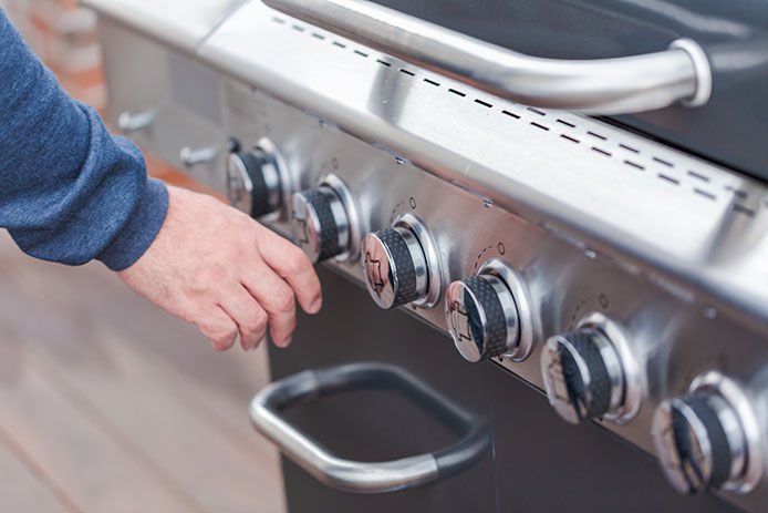 A person in a blue long-sleeved shirt reaches for the knob on the front of their stainless-steel gas grill. 