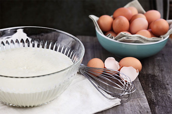 A bowl of waffle batter, a whisk and a bowl of eggs on a wooden tabletop