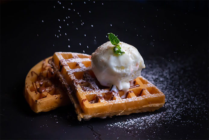 A couple of waffles with ice cream and chocolate drizzled on top and powdered sugar being sprinkled on top