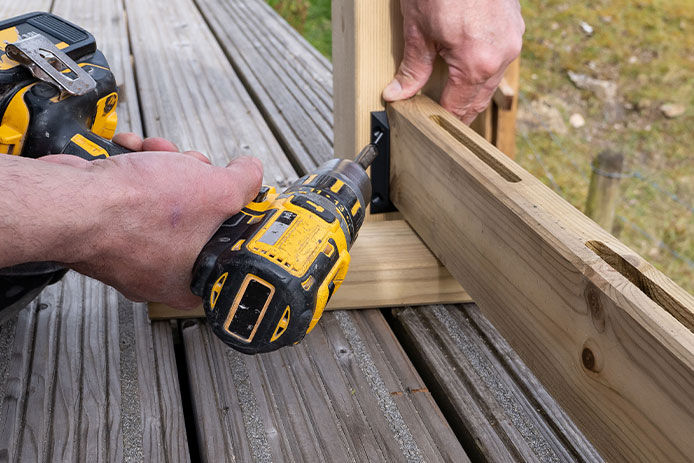 A carpenter working outdoors, building wooden decking on the side of family home. Using a handheld cordless battery drill he is securing the wooden railing posts on the edge of the deck.