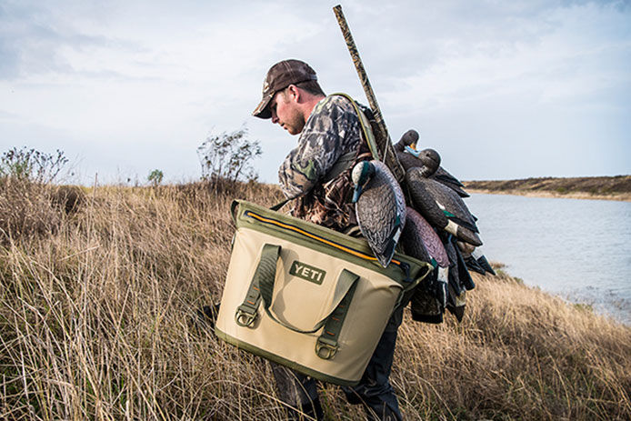 Man carrying duck decoys over his back with his rifle on one shoulder and a YETI cooler on the other