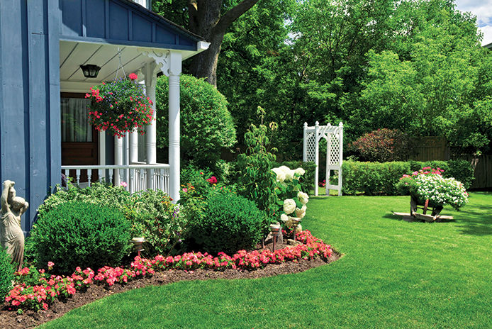 Front yard of a blue house with a white porch, adorned with a hanging basket. The yard is beautifully landscaped with red flowers and bushes, creating a welcoming atmosphere.
