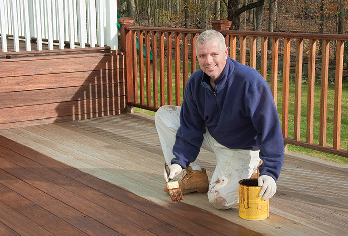 A grey-haired man in a navy-blue shirt and light-colored pants wears gloves and applies stain to a wooden deck. The stain is in a yellow-colored can, and the man uses a brush to apply the stain following the woodgrain of the deck.