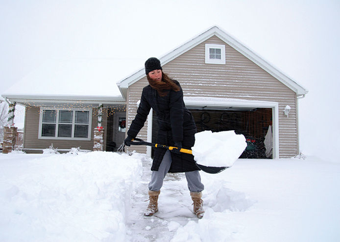 Woman scooping up a big pile of snow in her driveway wearing a long, black winter coat and hat