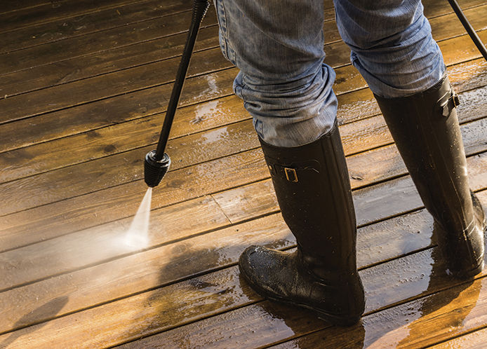 A person wearing jeans and black rubber rain boots uses a power washer to spray and clean the surface of a wooden deck.