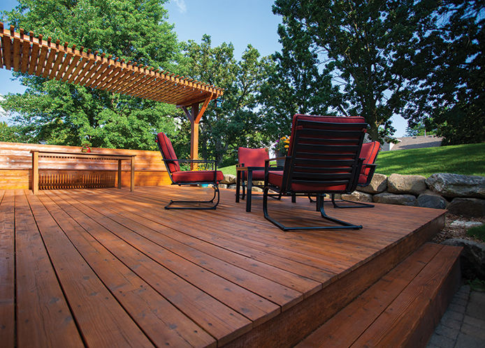 The image shows a stained wooden deck on a sunny day. The deck features four red patio chairs grouped around a table. 