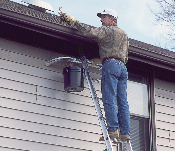 Man on a laddle with a bucket pullling leaves out of the gutter while wearing work gloves