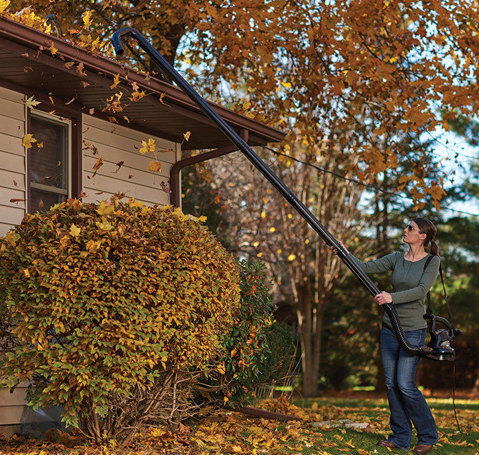 Woman using a leaf blower with extension to reach gutter to blow leaves out
