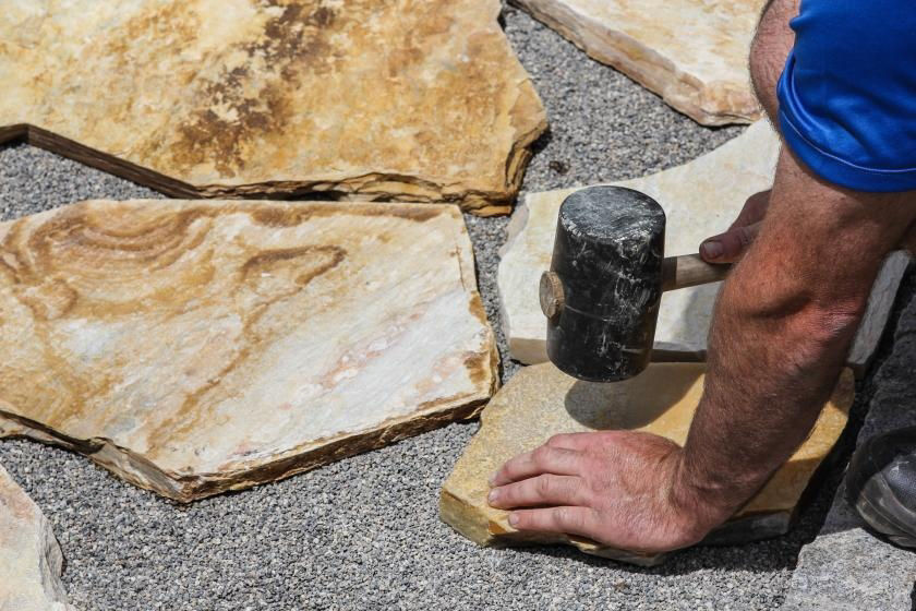 Man wearing a blue shirt using a rubber mallet to pound down a piece of flagstone into the pea gravel