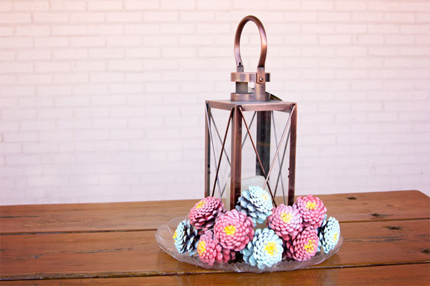 Final product. Zinnia painted pinecones around a wire lantern sitting on a wooden table 
