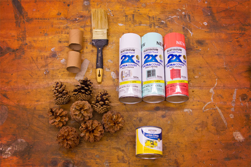 Painted Pinecone supplies. 3 cans of spray paint, a paint brush, a toilet paper roll, yellow acrylic paint and pinecones 