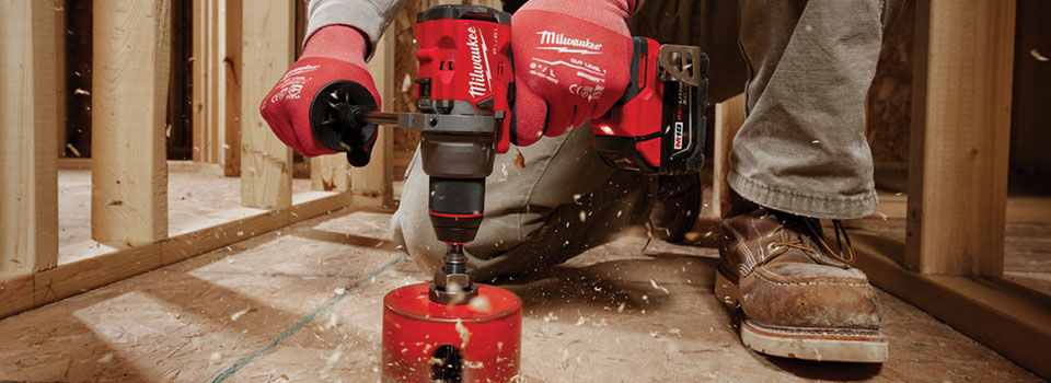 Worker using a cordless power drill with a hole saw attachment to cut a circular opening in a wooden floor during construction