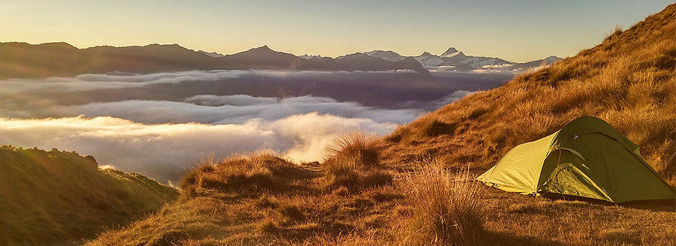 A green tent pitched on the side of a moutain horizon facing out into a morning sunrise with white fluffy clouds on the horizon.