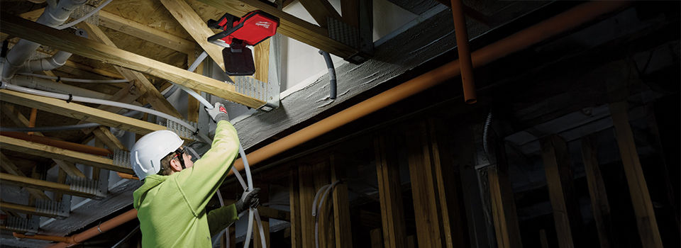 A man using a cordless work light to install electrical wire into the ceiling