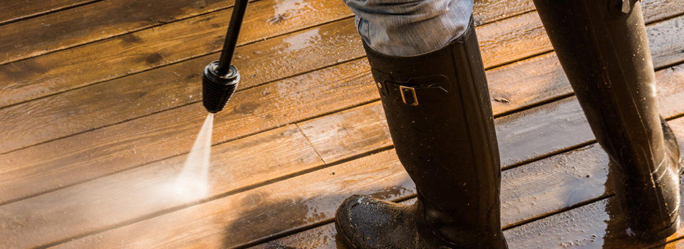A man wearing black rubber boots power washing a wooden deck