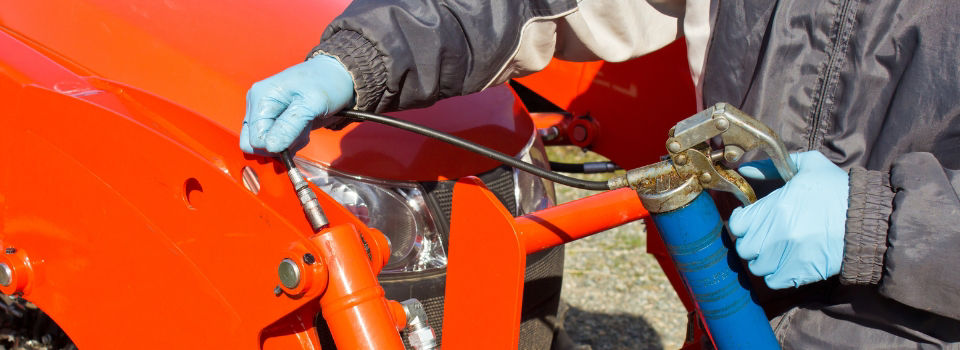 Person wearing gloves applies grease to an orange tractor using a grease gun