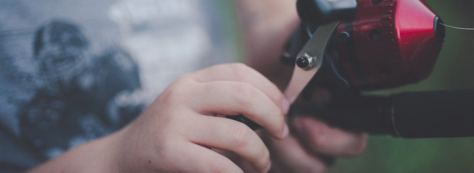 A young person in a grey tank top is holding a red and black fishing reel in their hands and turning the handle. 