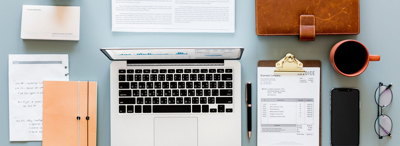 A top down view of an office desk with a laptop, clipboard, stack of papers, coffee cup, glasses, phone, and a brief case