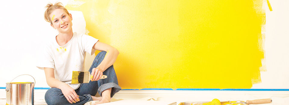 A young woman sitting on a wooden floor covered by a drop cloth surrounded by painting supplies and a bright yellow wall behind her
