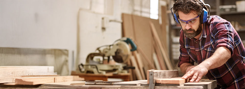 Skilled carpenter cutting a piece of wood in his woodwork workshop, using a circular saw, and wearing safety googles and earmuffs, with other machinery in the background