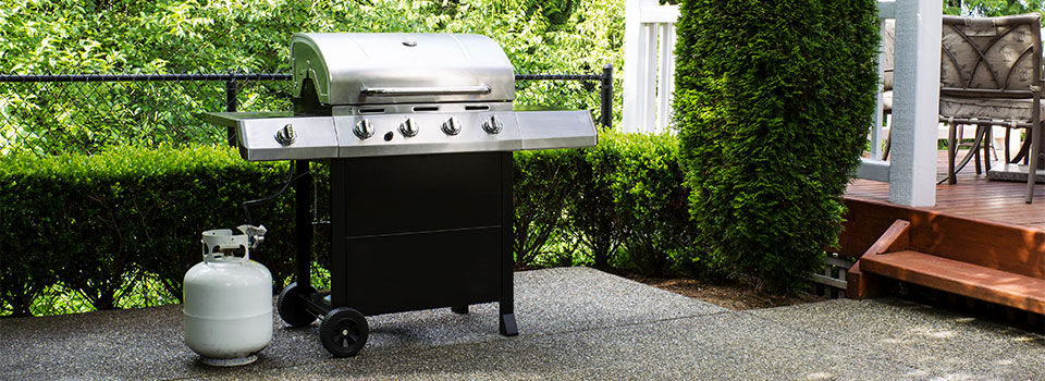 Stainless steel BBQ grill sitting on a level patio with white propane tank to the left and green shrubbery in the background