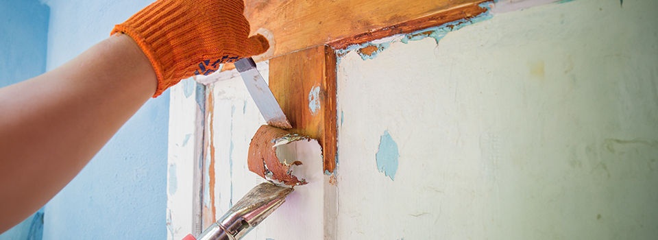 Person wearing orange work gloves using a heat gun and paint scraper to remove old paint off a wooden door