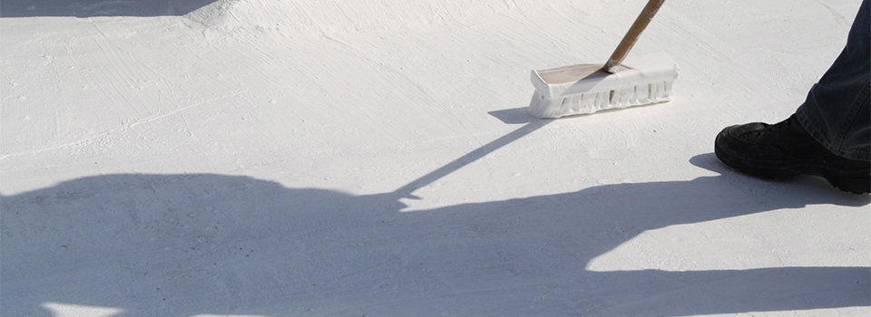 A man wearing black boot using a push broom to spread white roof coating over a rooftop