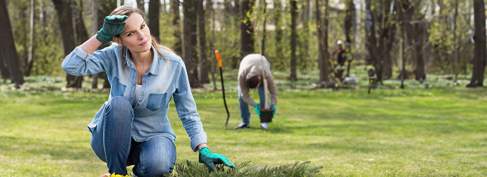 A brunette woman in a denim shirt and pants wipes her brow with a green-gloved hand as she crouches near a bush outside. In the background, a man holds a bucket and bends over next to a black and orange-handled yard tool. 