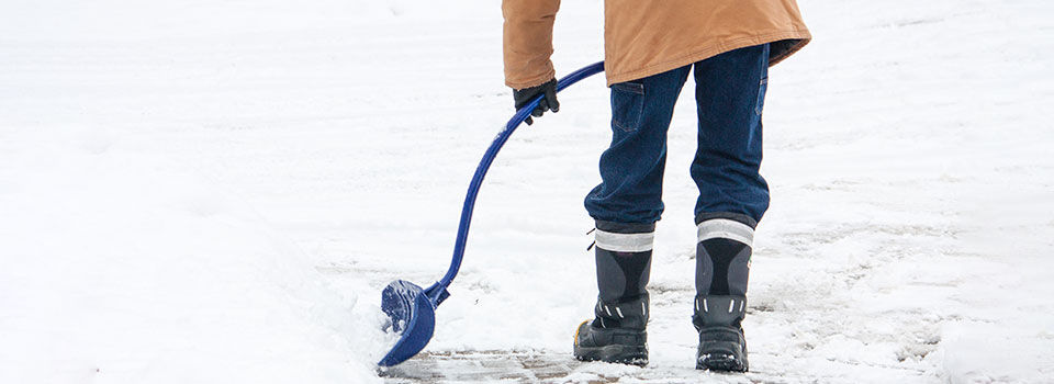 Person wearing black snow boots, jeans, and a tan winter coat shoveling snow from the driveway