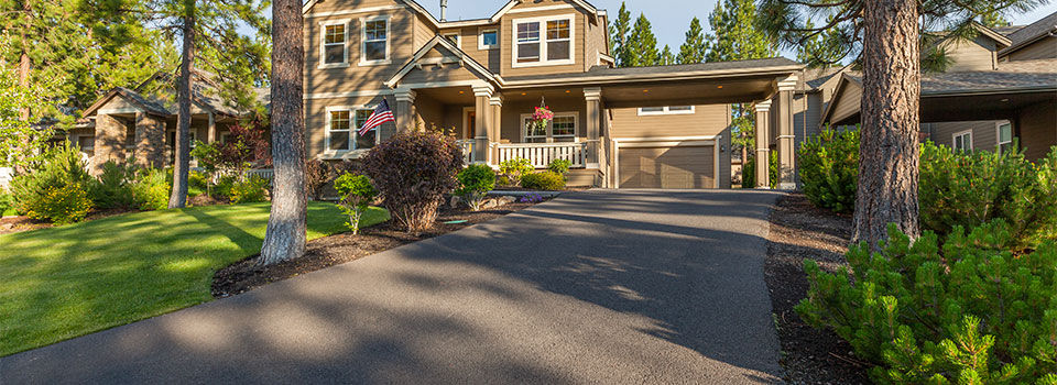 A large brown two-story house sits at the end of a long, freshly paved driveway. The driveway is dark grey and surrounded by green leafy trees and shrubs.