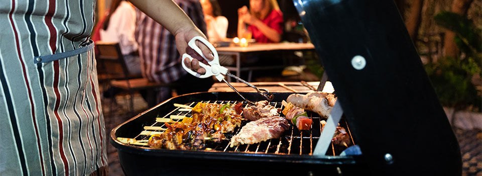 Person wearing an apron using tongs to flip meat on the grill with company in the background