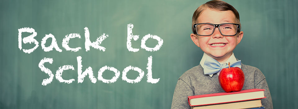 A young boy wearing thick rimmed glasses and holding a stack of books leans up against a chalkboard that reads back to school