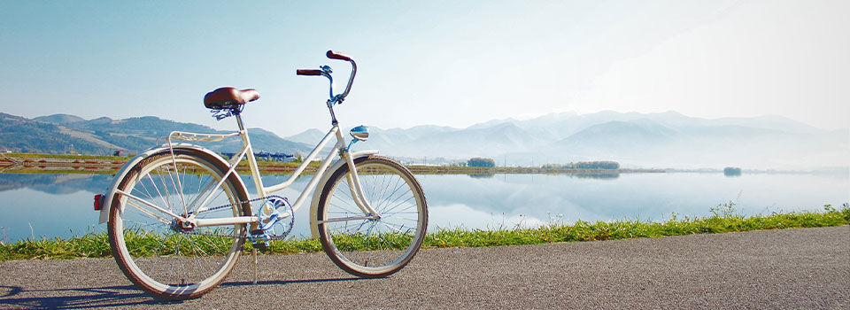 A blue and white cruiser bike is seen on a bike path with a beautiful blue lake and mountain range in the background. The bike has a comfortable-looking seat, white-walled tires, and a classic frame design. The background features a clear blue lake, with ripples visible on the surface, and a majestic mountain range rising behind it. 