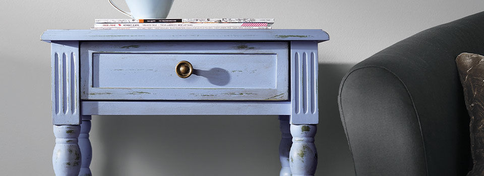  A matte sea blue end table with a black knob gets its distinct finish from chalk paint. The farmhouse-style table sits against a white wall.