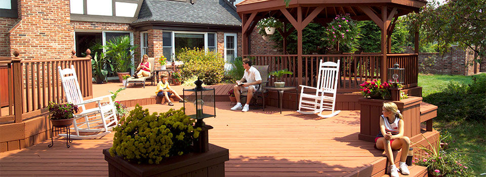 A family enjoys their wooden deck and backyard gazebo on a sunny summer day. The deck includes two white rocking chairs with a brown brick house in the background. 