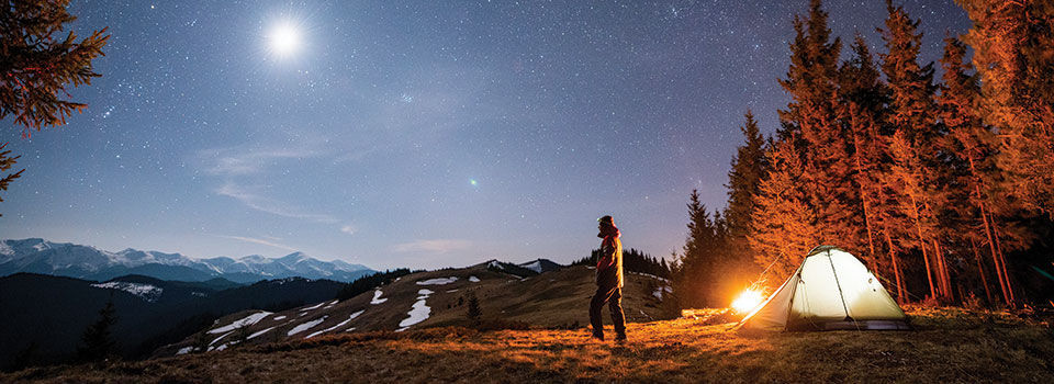 Man wearing a jacket looking up at the night sky with a fire going over by his tent