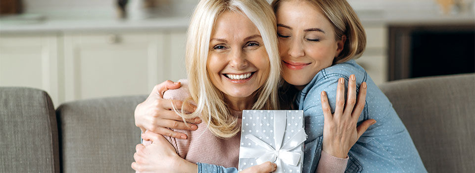 Surprise for mother's day or birthday. Loving young adult daughter giving a present to her beloved middle-aged caucasian mom sitting on the sofa in the living room