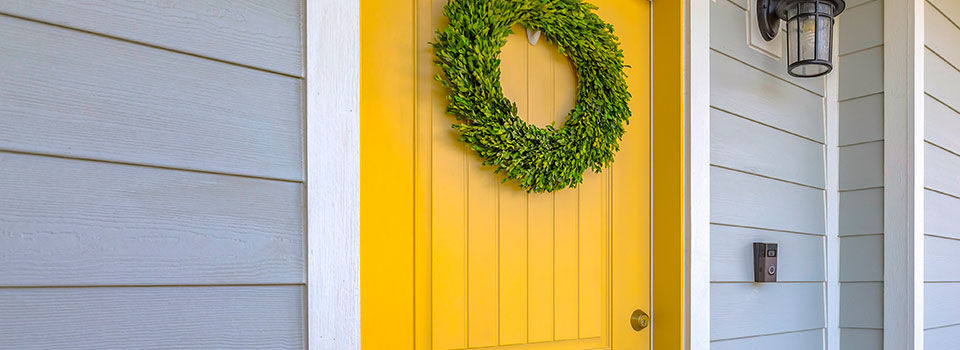 Residential home with a brightly yellow painted from door. The door has a green wreath and white trim. The house is also a light blue gray color with an light fixture accent above the doorbell.