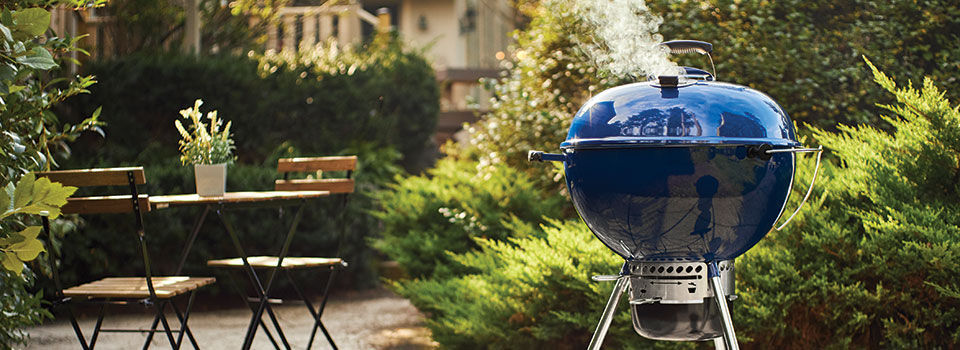 A blue charcoal grill sitting on a patio with a two chair chat set and green foilage surrounding