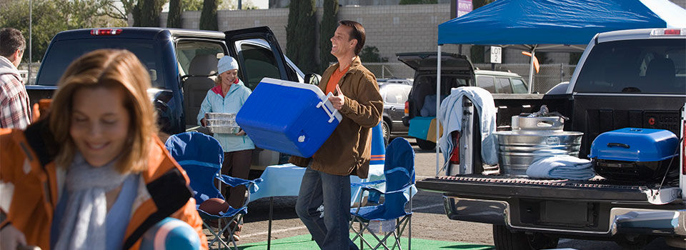 Man carrying a cooler from the back of a pickup truck in the midst of people tailgating
